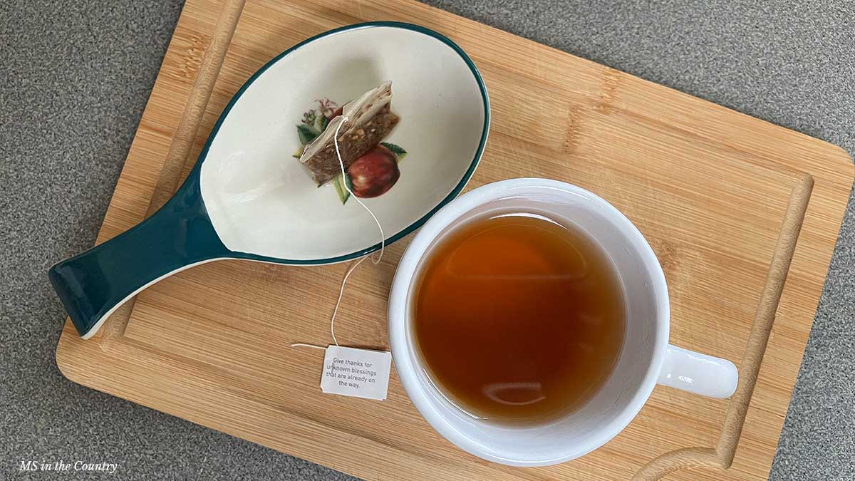 A white mug of tea and used tea bag on a spoon rest with apple design, styled on a wooden board – part of the Nourish and Heal Library