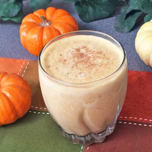 Pumpkin Pie Smoothie in a glass topped with nutmeg, surrounded by small pumpkins on a fall-colored napkin.