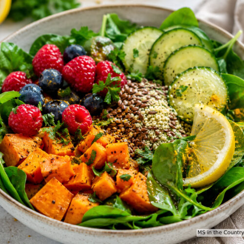 A gut healthy bowl with spinach, roasted sweet potato cubes, raspberries, blueberries, flaxseeds, hemp seeds, cucumber slices, and lemon, topped with fresh herbs