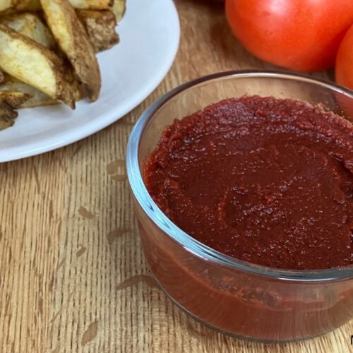 Homemade ketchup in a glass bowl served with potato wedges and fresh tomatoes on a wooden cutting board