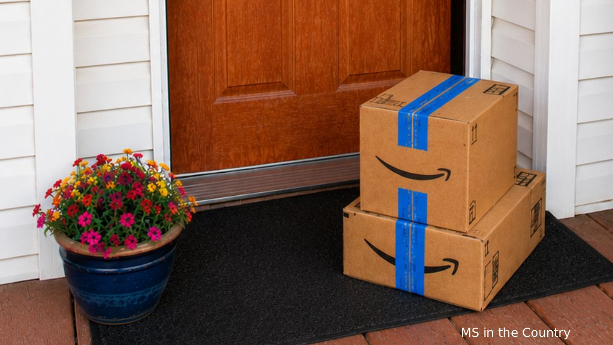 Amazon Prime Day 2026 packages stacked on a front porch beside colorful flowers and a rustic firewood stack.