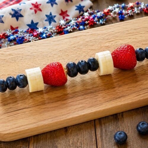 Red, white, and blue 4th of July fruit skewer with blueberries, banana, and strawberries on a wood cutting board with patriotic star garland