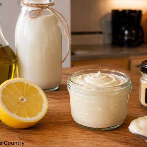 Homemade mayonnaise without eggs in a glass jar surrounded by olive oil, almond milk, lemon, sea salt, and mustard powder