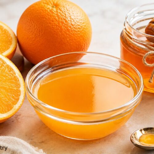 Honey orange glaze in a glass bowl with fresh oranges, a jar of raw honey with a honey dipper, and a vintage spoon