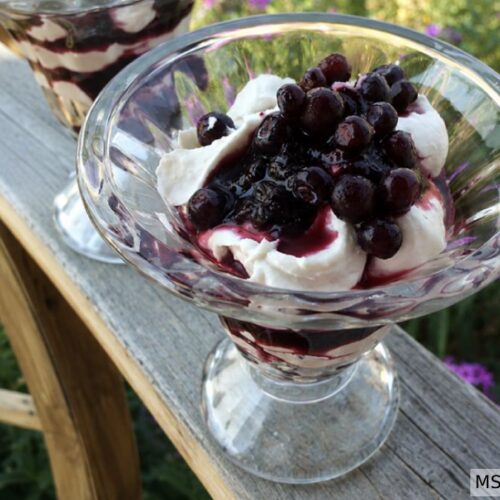 Huckleberry coconut whipped cream layered in crystal glasses on a wooden porch railing with purple flowers and wagon wheel